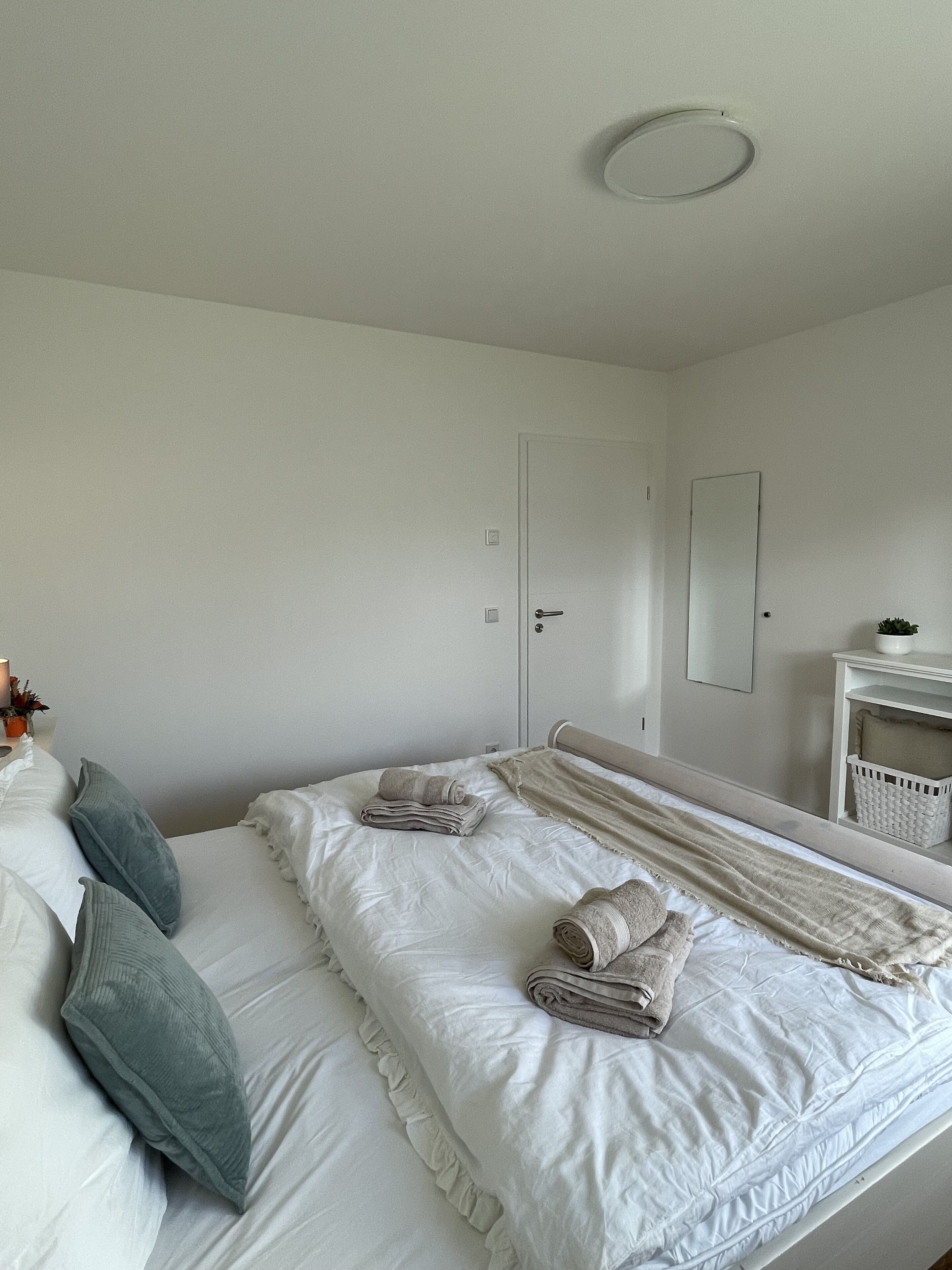 A neatly made bed with pillows and folded towels, a bedside table with a basket, and a mirror on the wall.
