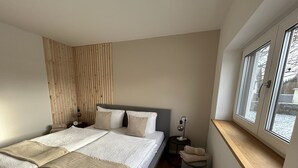 Ein modernes Schlafzimmer mit einem großen Bett, einem Holzkopfende, einem Nachttisch mit Lampe, einem kleinen Hocker und einem Fenster mit Blick.