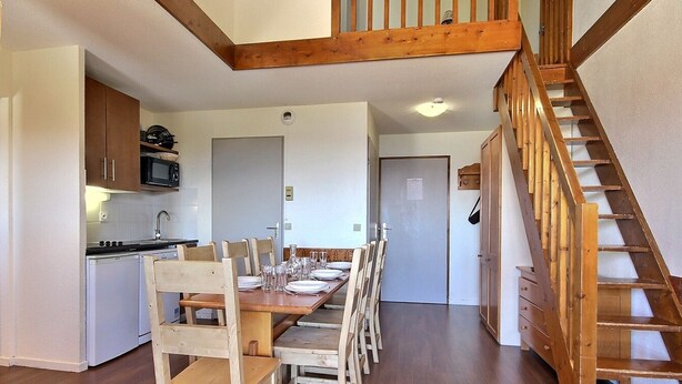 A dining area with a wooden table set for six, wooden chairs, a kitchen with a microwave, and a wooden staircase. - Spacious duplex for 5 at the foot of the slopes (Plagne Soleil)