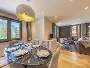 A modern dining area with a round table set for six, a sofa, and a TV in a living area with large windows.