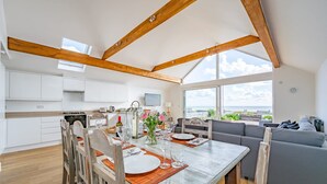 Een moderne keuken met witte kasten, een eetgedeelte met een houten tafel voor vier personen, en een grijze bank in een woonruimte met grote ramen.