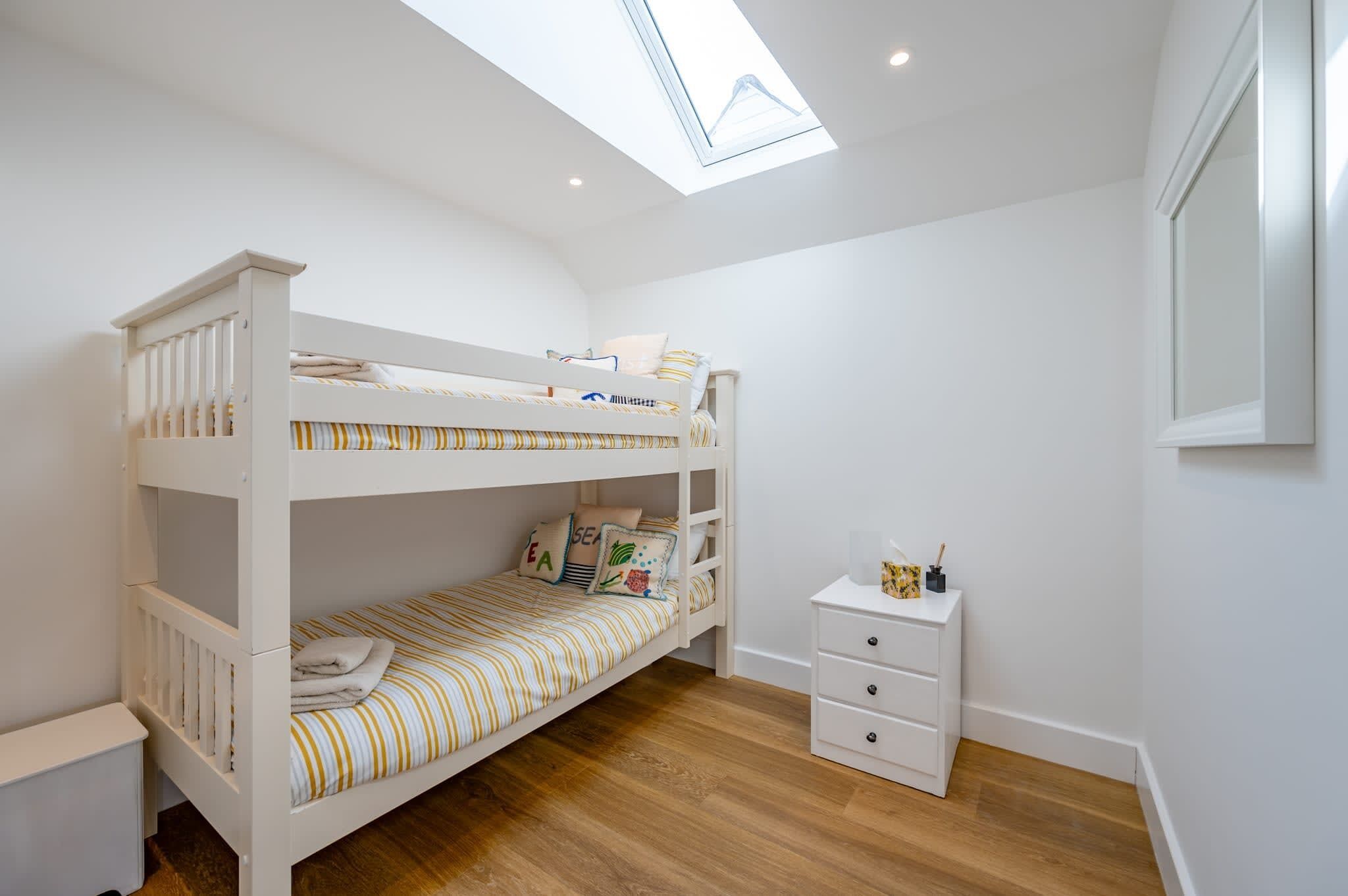 Une chambre avec un lit superposé, une lucarne, une petite commode blanche et une fenêtre.