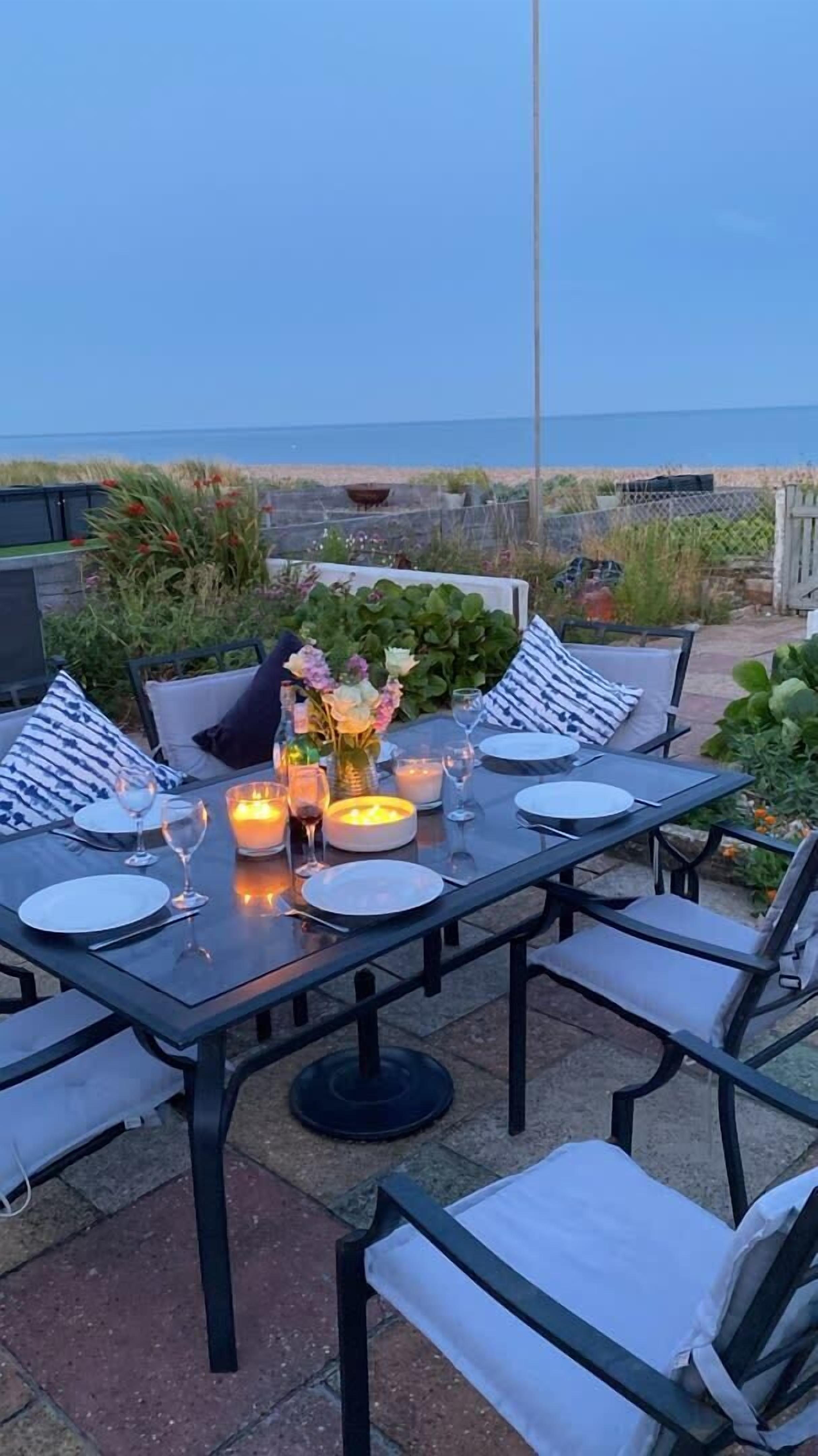 An outdoor dining area with a table set for four, surrounded by chairs with cushions. There are candles on the table, and a view of the ocean in the background.