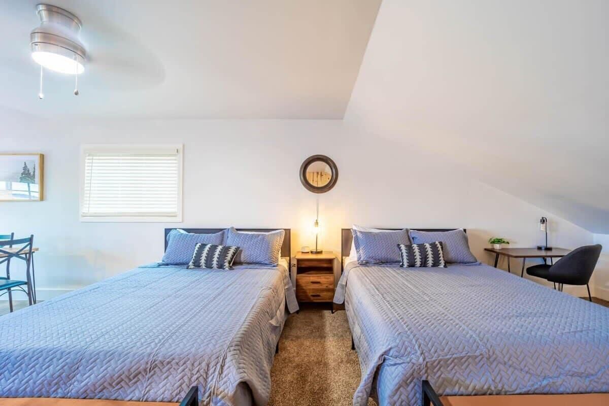 A bedroom with two beds, a small table, a chair, a wall-mounted lamp, and a ceiling light fixture.