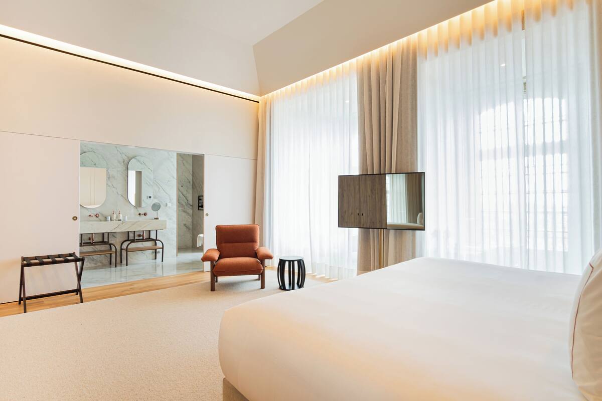 Una habitación de hotel moderna con una cama grande, un televisor, una zona de estar con una silla y una mesita auxiliar, y un espejo amplio.