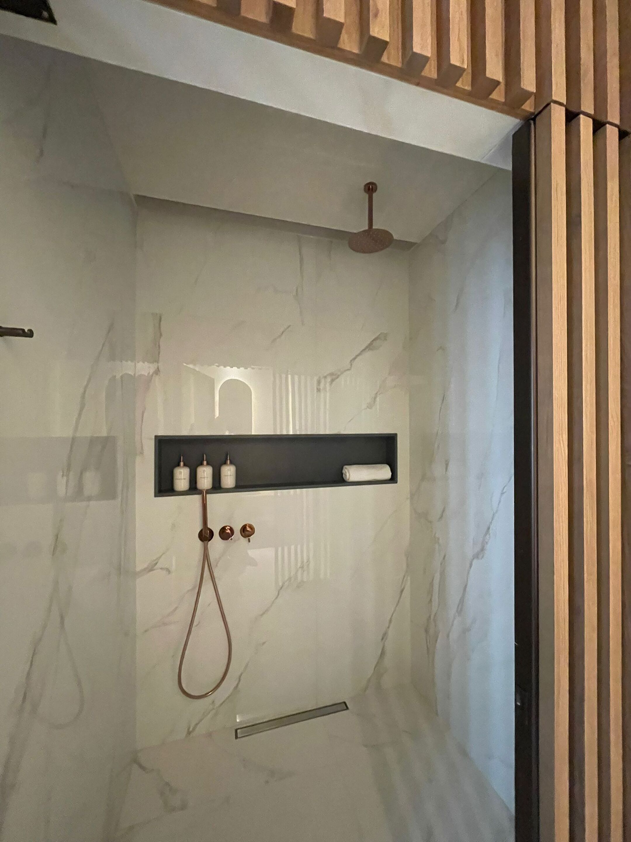 A modern bathroom with a marble wall, a shower with a handheld showerhead, and a shelf with toiletries.