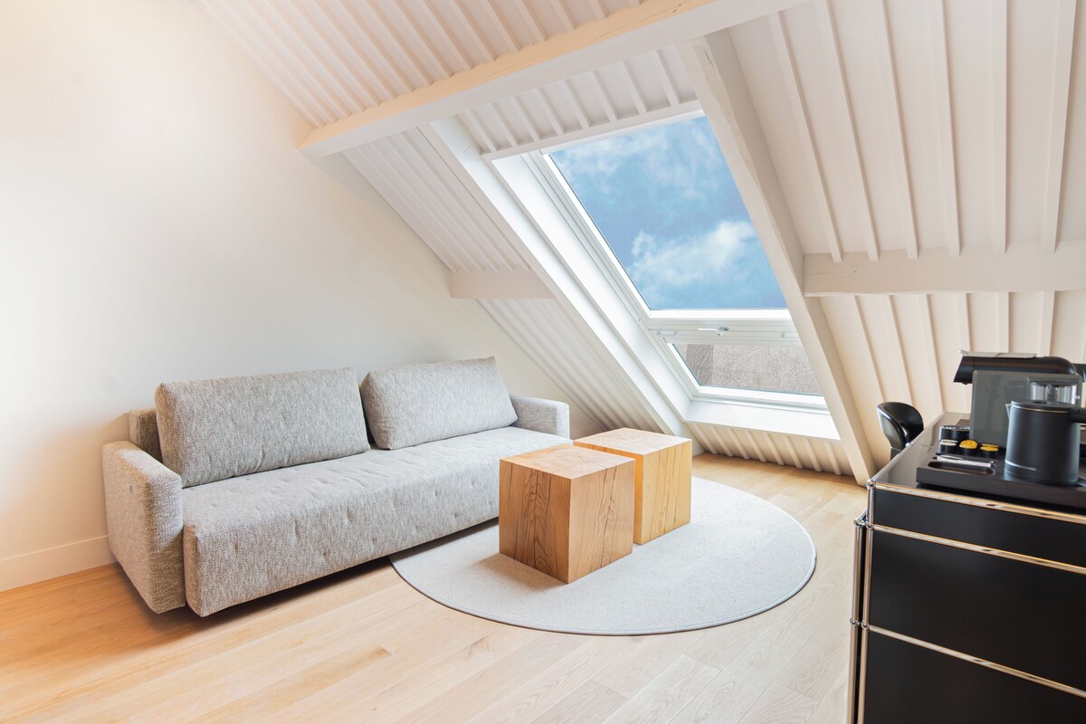 Ein modernes Schlafzimmer im Loft-Stil mit schrägen Wänden, einem grauen Sofa, einem Holztisch und einer schwarzen Kommode mit Wasserkocher.
