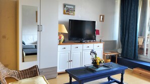 A room with a television on a wooden cabinet, a wicker chair, a small table with a flower vase, and a window with curtains. - Studio Glovettes 35m², Perfect for Ski & Hiking (Villard-de-Lans)