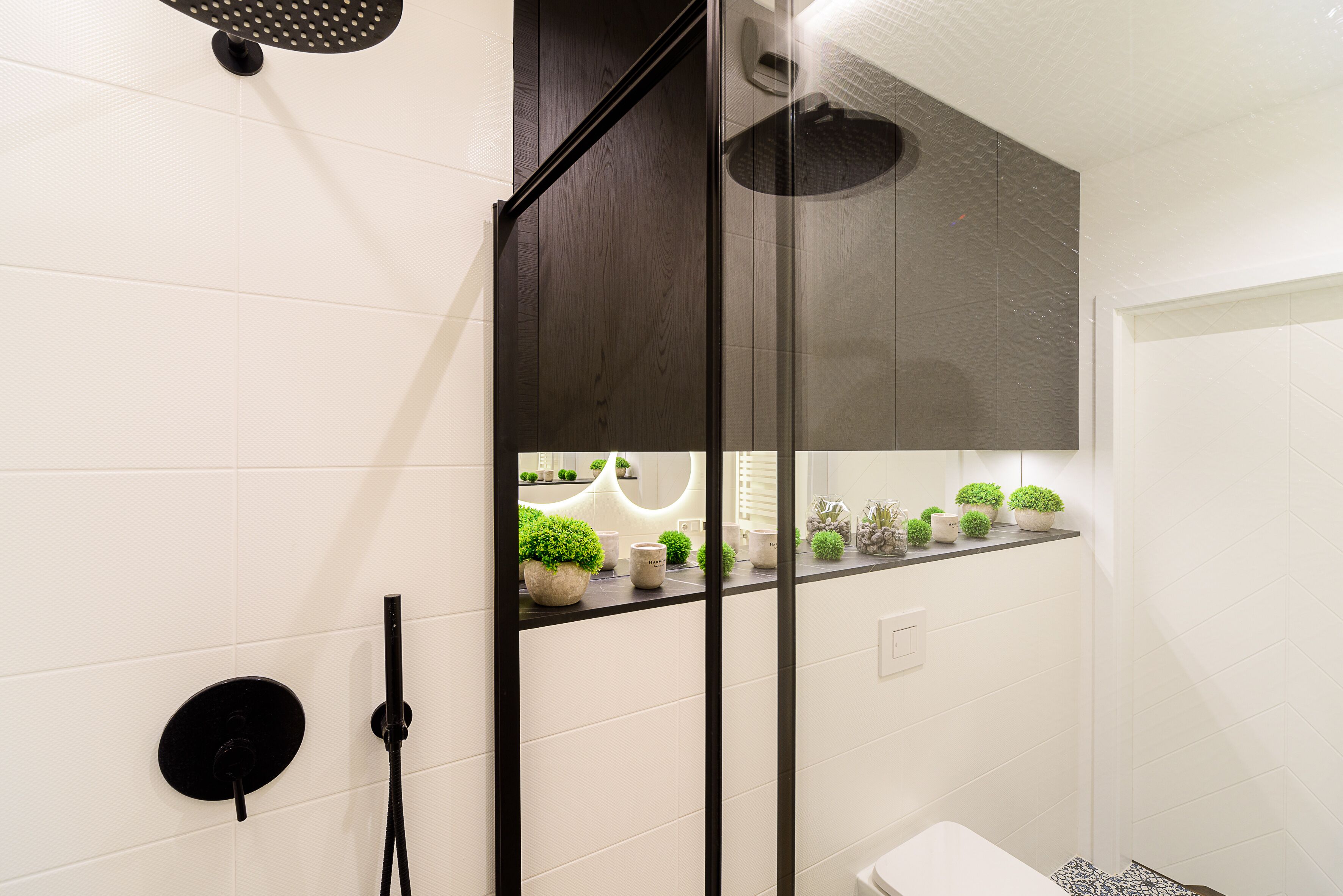 Ein modernes Badezimmer mit weißen Fliesen, einer Duschkabine aus Glas, einer schwarzen Toilette und einem Regal mit Topfpflanzen.