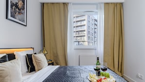 Habitación de hotel con cama, ventana con cortinas, un cuadro en la pared y una bandeja con queso, uvas y copas de vino.