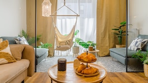 Una sala de estar acogedora con un sofá, una silla, una mesita de centro con croissants y un lámpara colgante.