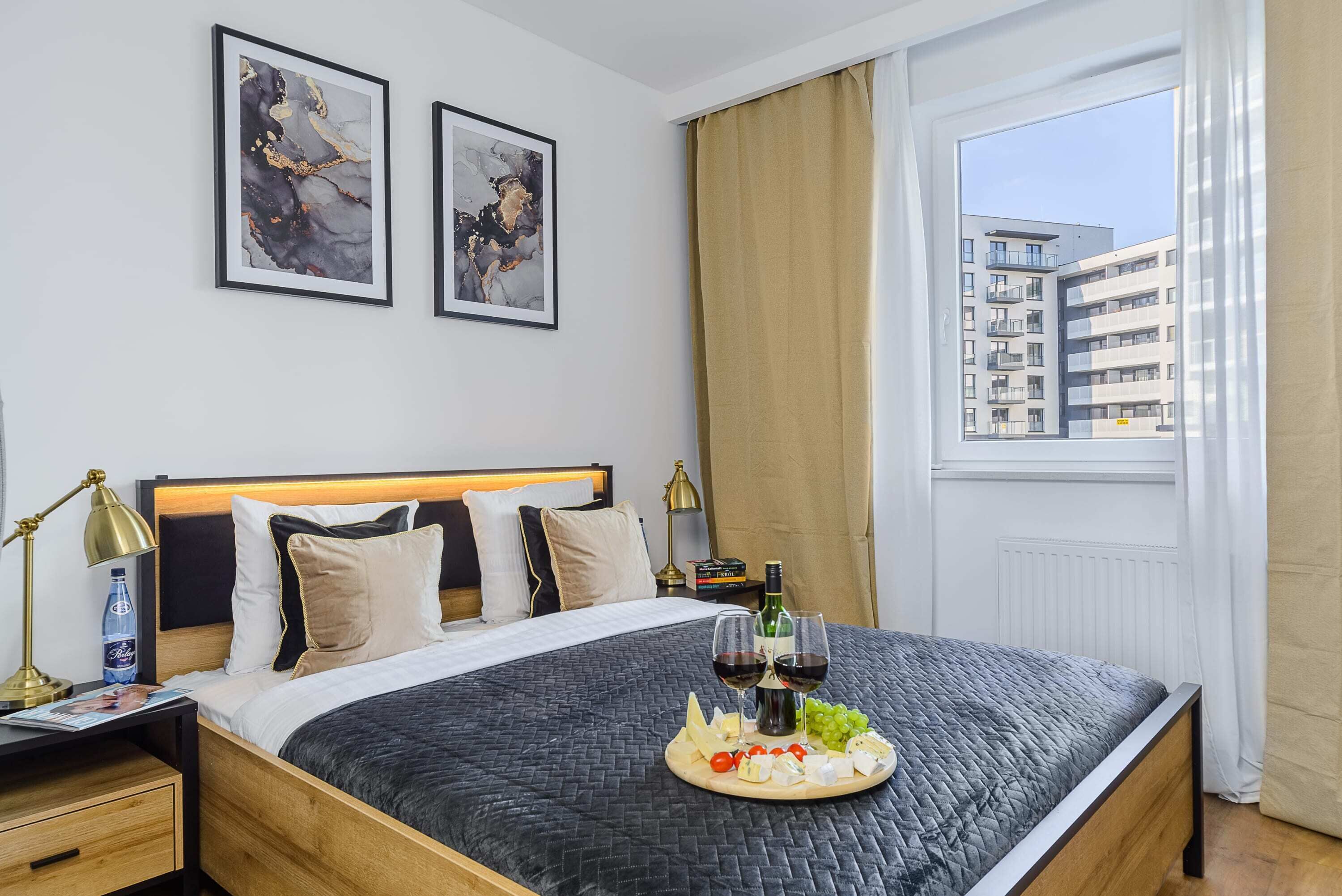 A hotel room with a bed, a bedside table with a lamp, a tray with food and wine, and a window with a view of buildings.