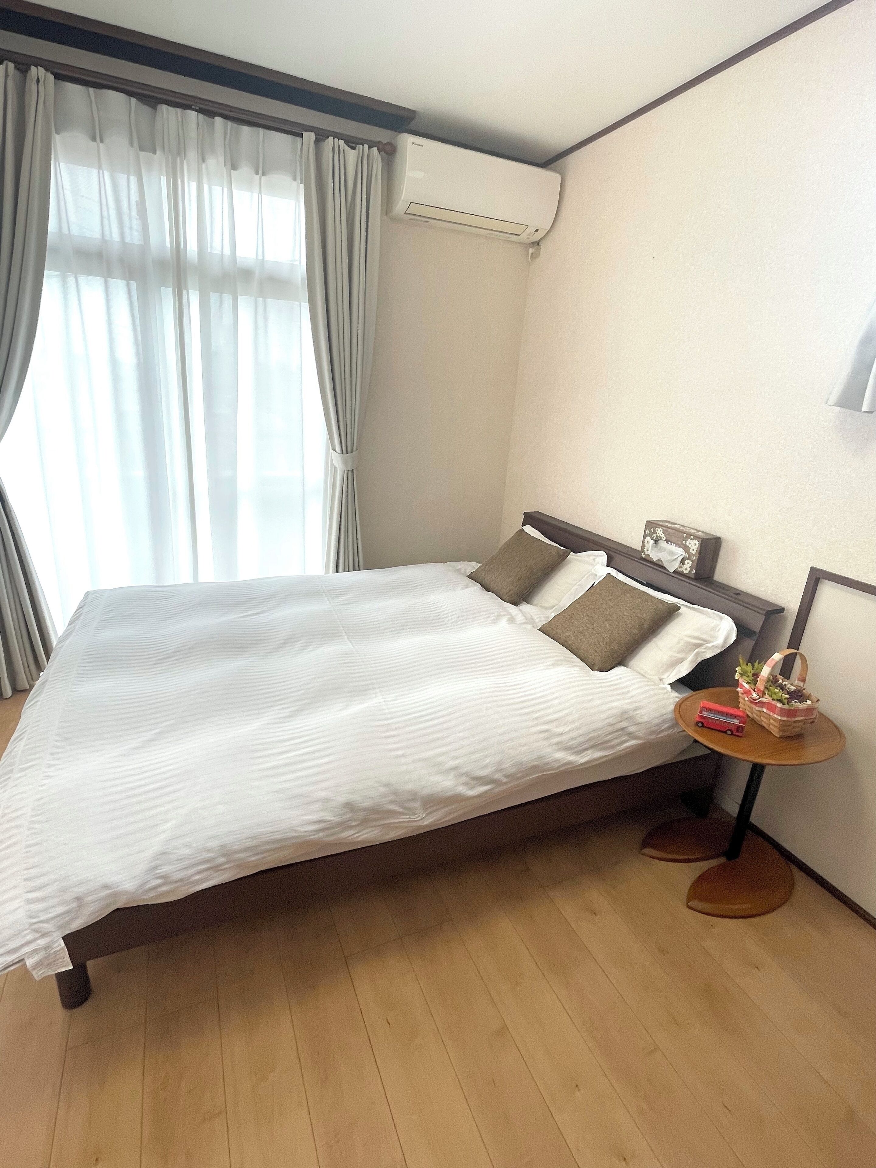 A neatly made bed with white linens and two pillows, a bedside table with a basket and a red object, and an air conditioning unit mounted on the wall.