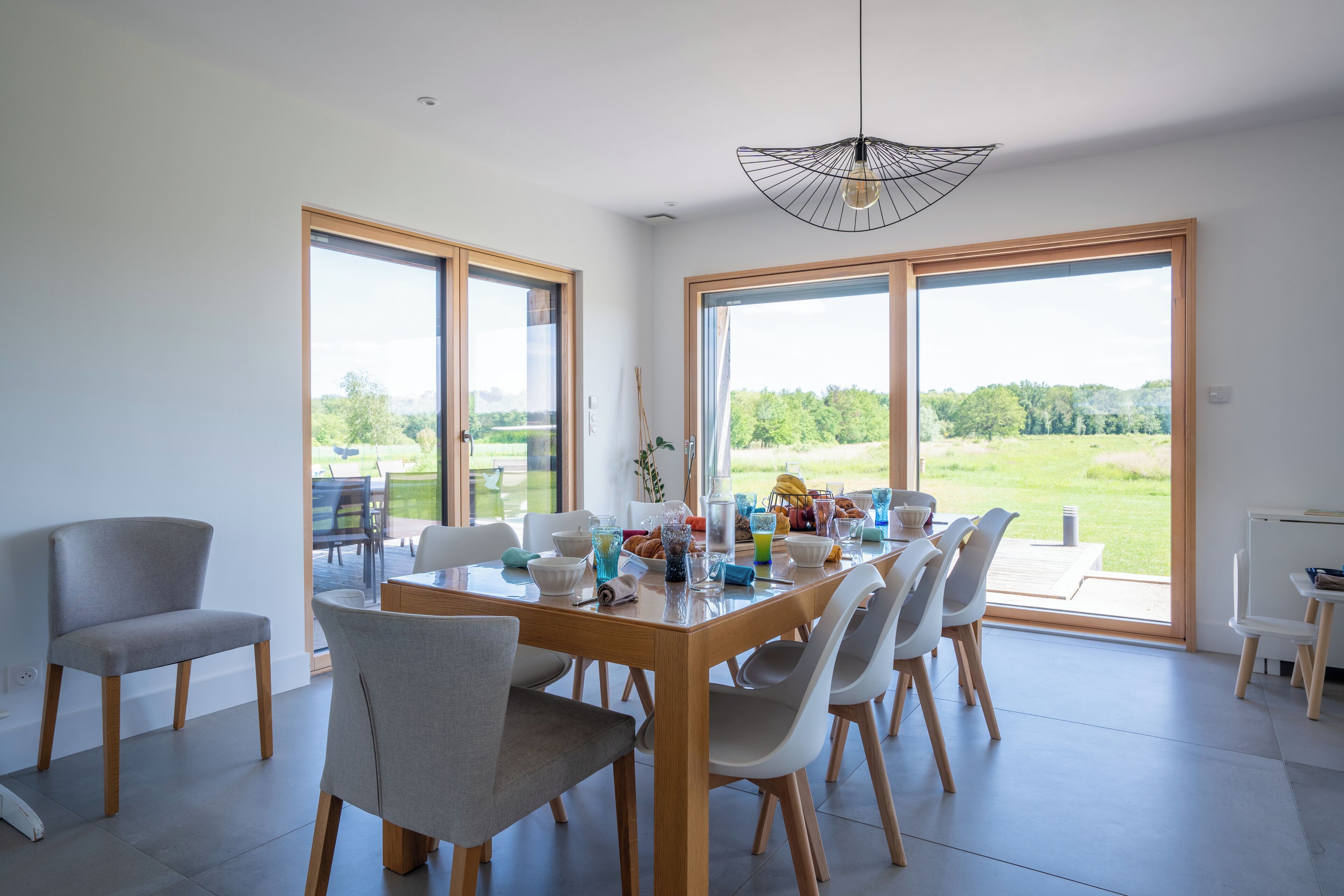 Un vaste espace dédié à la salle à manger, avec une table et des chaises en bois, de grandes fenêtres offrant une vue sur l’extérieur et un luminaire suspendu moderne.