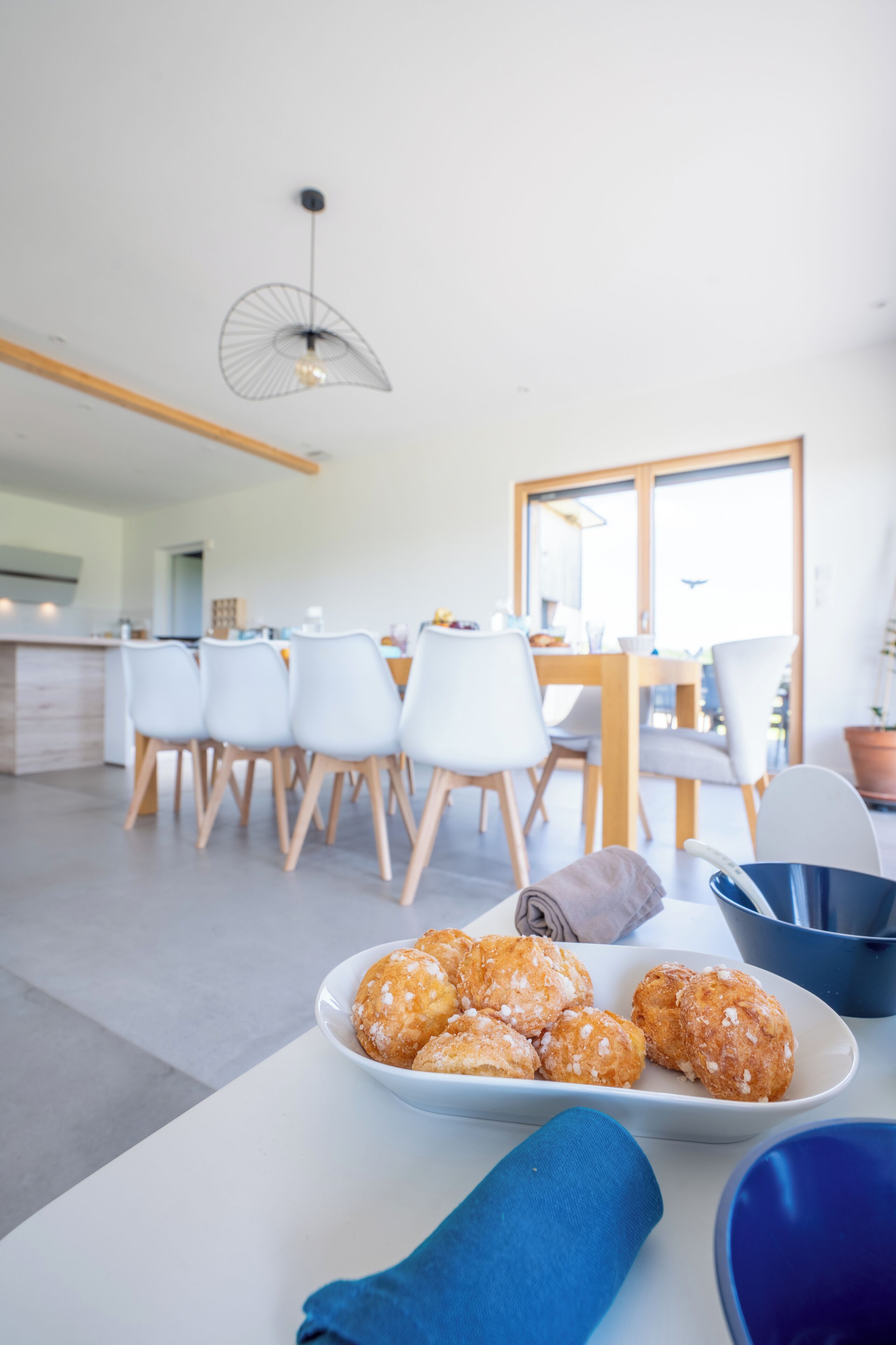 A modern kitchen with a dining area, featuring a white table with a plate of pastries, a blue napkin, and a blue bowl.