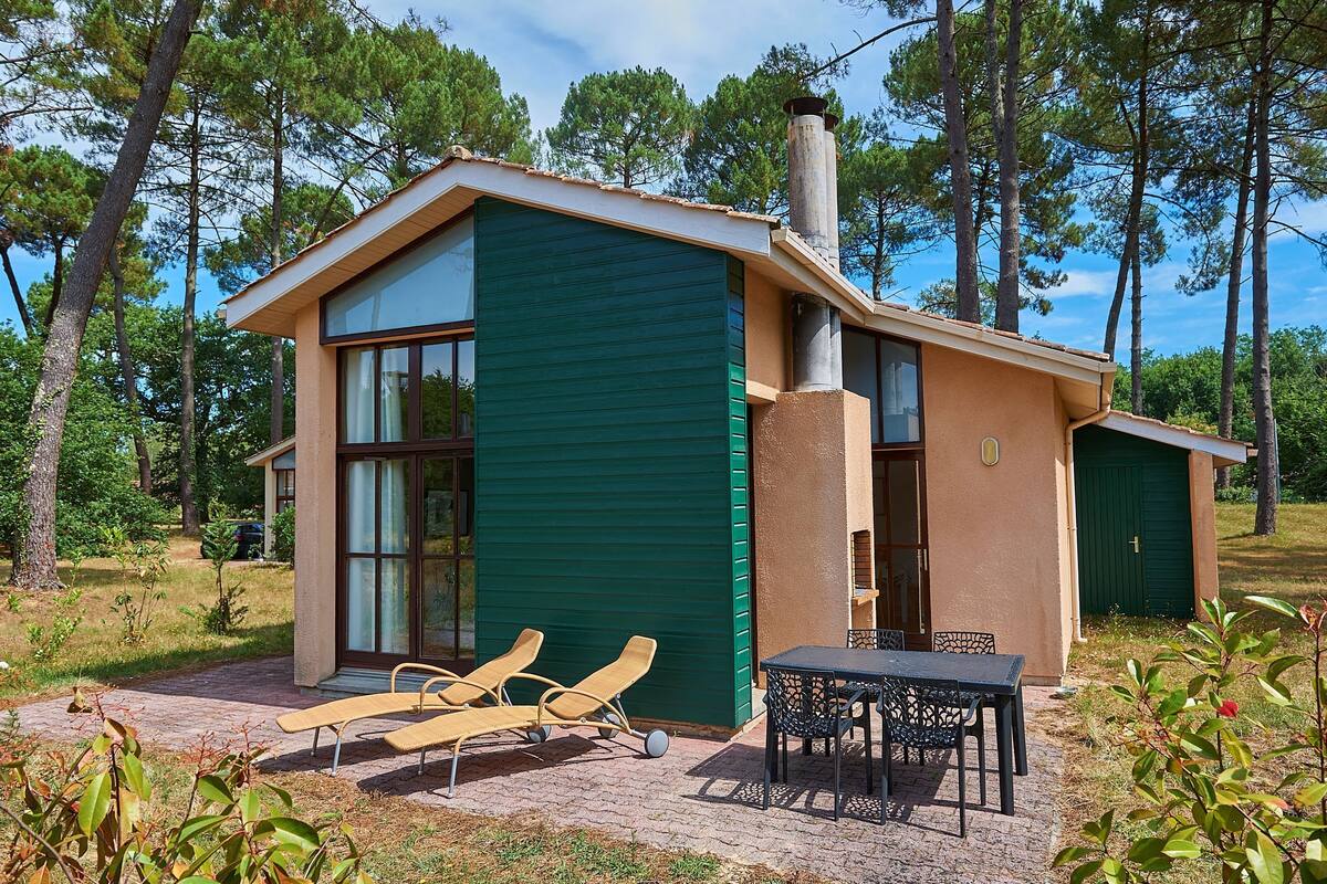 Une maison moderne avec une façade verdoyante, de grandes fenêtres et un patio aménagé avec du mobilier d’extérieur.