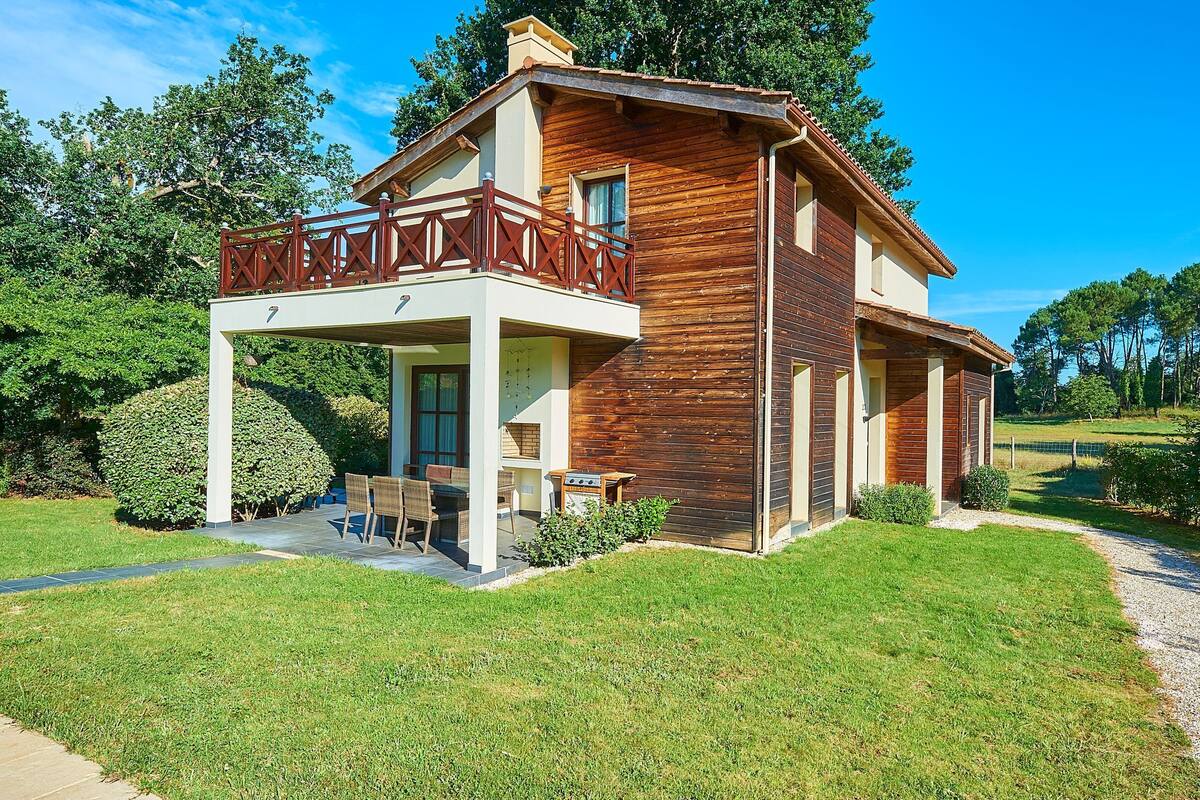 Une maison en bois à deux étages, dotée d’un balcon, d’une terrasse avec des meubles d’extérieur et d’une pelouse bien entretenue.