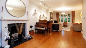 A living room with a fireplace, a grand piano, a round mirror, and a cozy seating area. - Majestic Oasis, by Veeve (London)