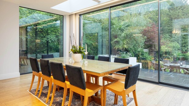 A dining area with a glass table and chairs, large windows, and a view of a lush garden. - Majestic Oasis, by Veeve (London)