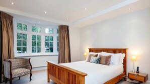 A bedroom with a large bed, a wooden nightstand, a chair, and a window with curtains. - Majestic Oasis, by Veeve (London)