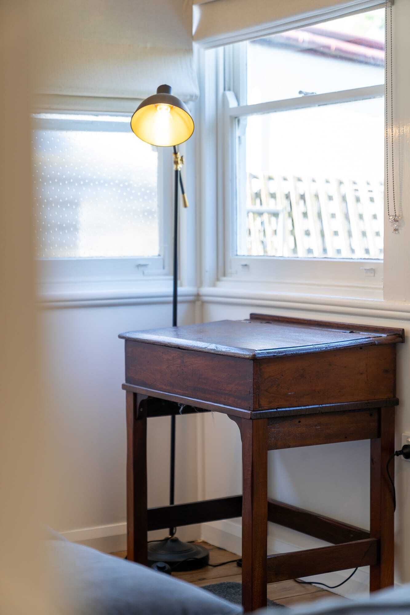 Een hoekzaak met een houten tafel, een vloerlamp en een raam met jaloezieën.