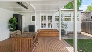 A covered patio with a wooden table and chairs, a small kitchen area, and a view of the backyard. - Johnno's Cottage - Prime Location - Sleeps 4 (Orange)