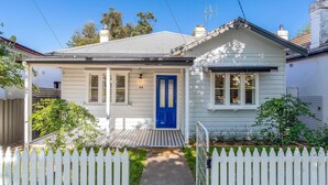A single-story house with a blue front door, white picket fence, and a small front yard. - Johnno's Cottage - Prime Location - Sleeps 4 (Orange)