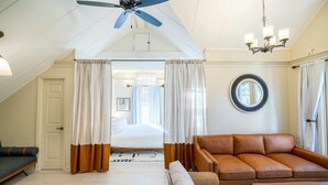 Amplia sala de estar con ventilador de techo, un sofá de cuero marrón, una mesa de centro de madera y vista al dormitorio a través de una puerta con cortinas.