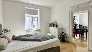A bedroom with a bed, bedside table, and a window. There is a dining area with a table and chairs visible through an open door. - 90 m2 - Space to Chill & Work & Rest (München)