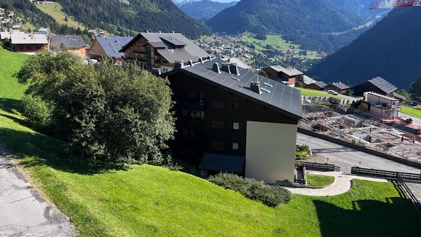 A mountain village with traditional wooden houses, a modern building with solar panels, and a clear view of the surrounding mountains.