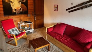A living room with a red sofa, a wooden chair with a red cushion, a small wooden table, and a large wall painting of a cow.
