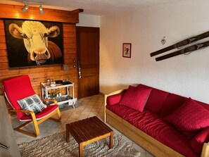 A living room with a red sofa, a wooden chair with a red cushion, a small wooden table, and a large wall painting of a cow.