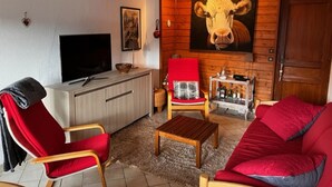 A living room with a TV, a red sofa, a red armchair, a wooden coffee table, and a wall-mounted painting of a cow.