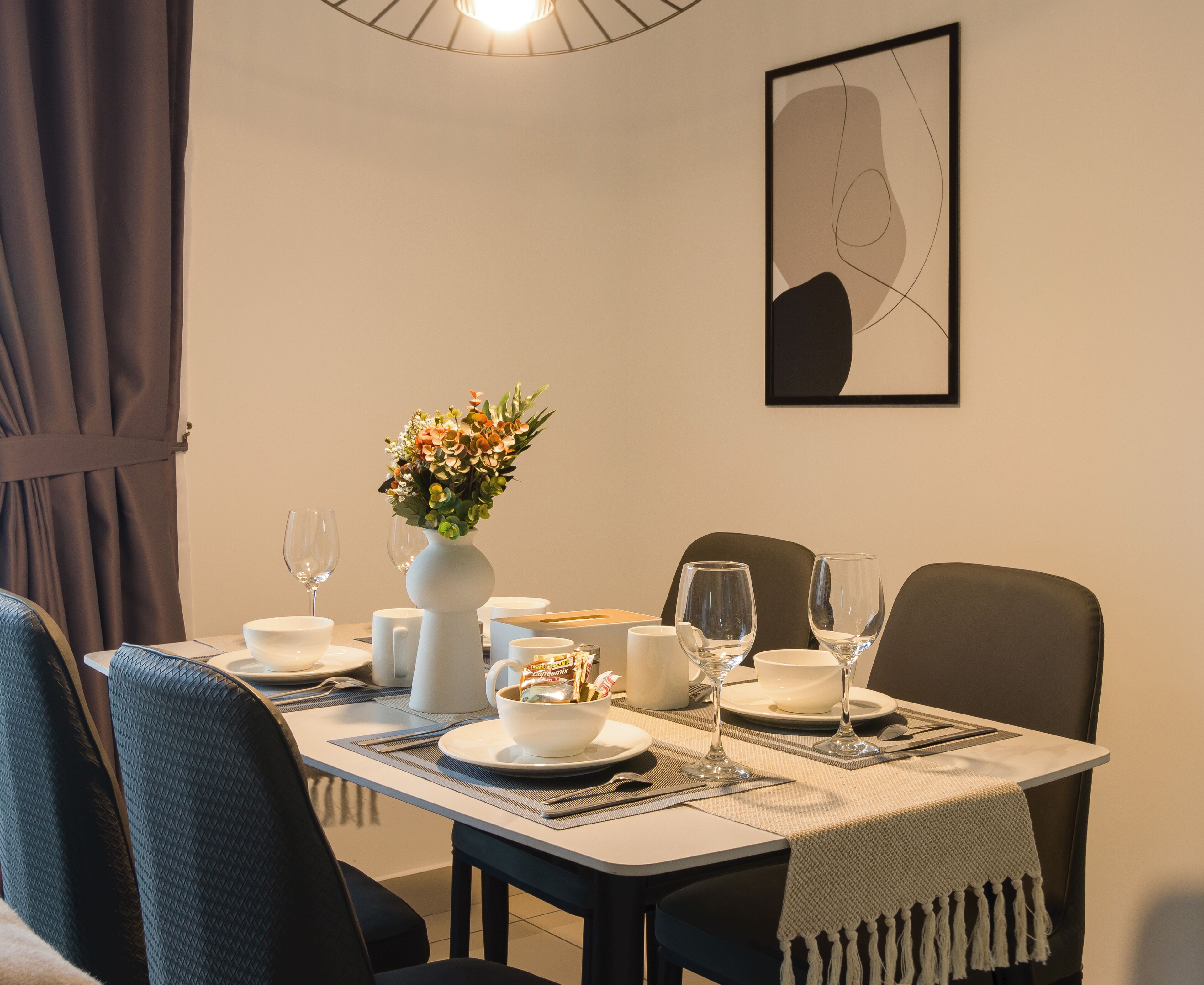 Une table à manger dressée avec de l’assiette, des bols et des verres blancs, un vase rempli de fleurs et une œuvre d’art abstraque encadrée accrochée au mur.