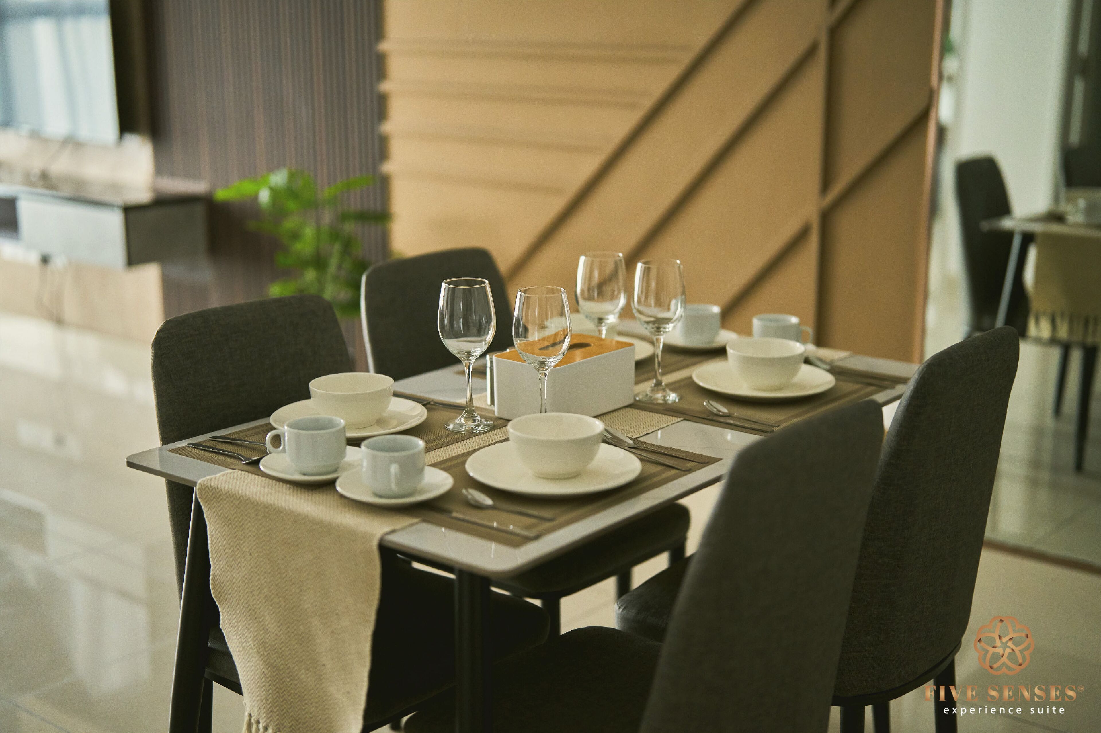 Une table à manger dressée avec de l’assiette, des bols et des verres à vin blancs, recouverte d’une nappe beige et entourée de chaises foncées.