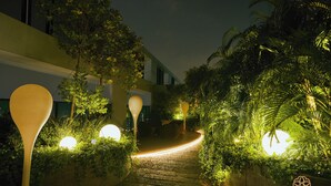 Un chemin bordé d’une végétation luxuriante et éclairé par des lampes sphériques, qui mène à un bâtiment au design moderne.