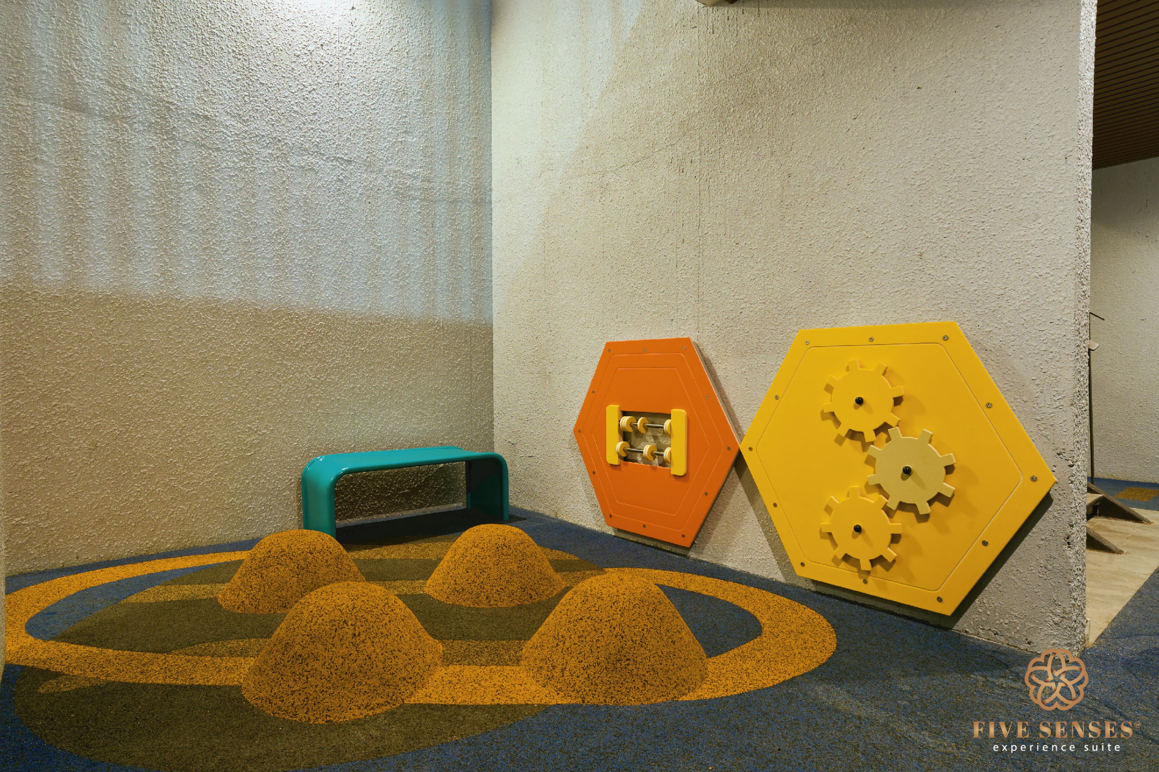 Un espace de jeux doté d’un sol souple, d’une structure de jeux bleue et orange, et d’un panneau de jeux jaune en forme d’hexagone.