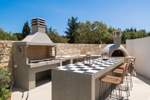 Una cocina al aire libre con pared de piedra, horno para pizzas y una mesa a cuadros.