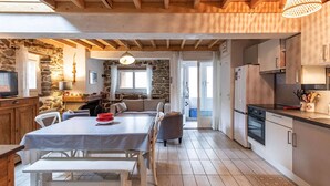 A cozy kitchen with a dining area, stone walls, wooden beams, and a view to the outside.