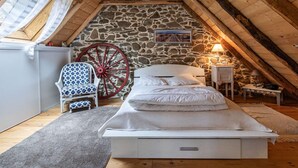 A cozy attic bedroom with a stone wall, a wooden ceiling, a bed, a rocking chair, and a red wagon wheel decoration.
