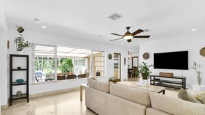 A spacious living room with a large window, a ceiling fan, and a flat-screen TV. - Bungalow 31 – Coral Ridge Oasis | Heated Pool, Private Escape, Just 6 Mins to the Beach! (Fort Lauderdale)