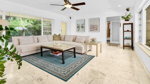 A spacious living room with a large sectional sofa, a coffee table, and a patterned rug. There is a ceiling fan, a small side table, and a bookshelf with decorative items. - Bungalow 31 – Coral Ridge Oasis | Heated Pool, Private Escape, Just 6 Mins to the Beach! (Fort Lauderdale)