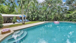 A swimming pool with a clear blue water, surrounded by palm trees and a covered area with chairs and a table. - Bungalow 31 – Coral Ridge Oasis | Heated Pool, Private Escape, Just 6 Mins to the Beach! (Fort Lauderdale)
