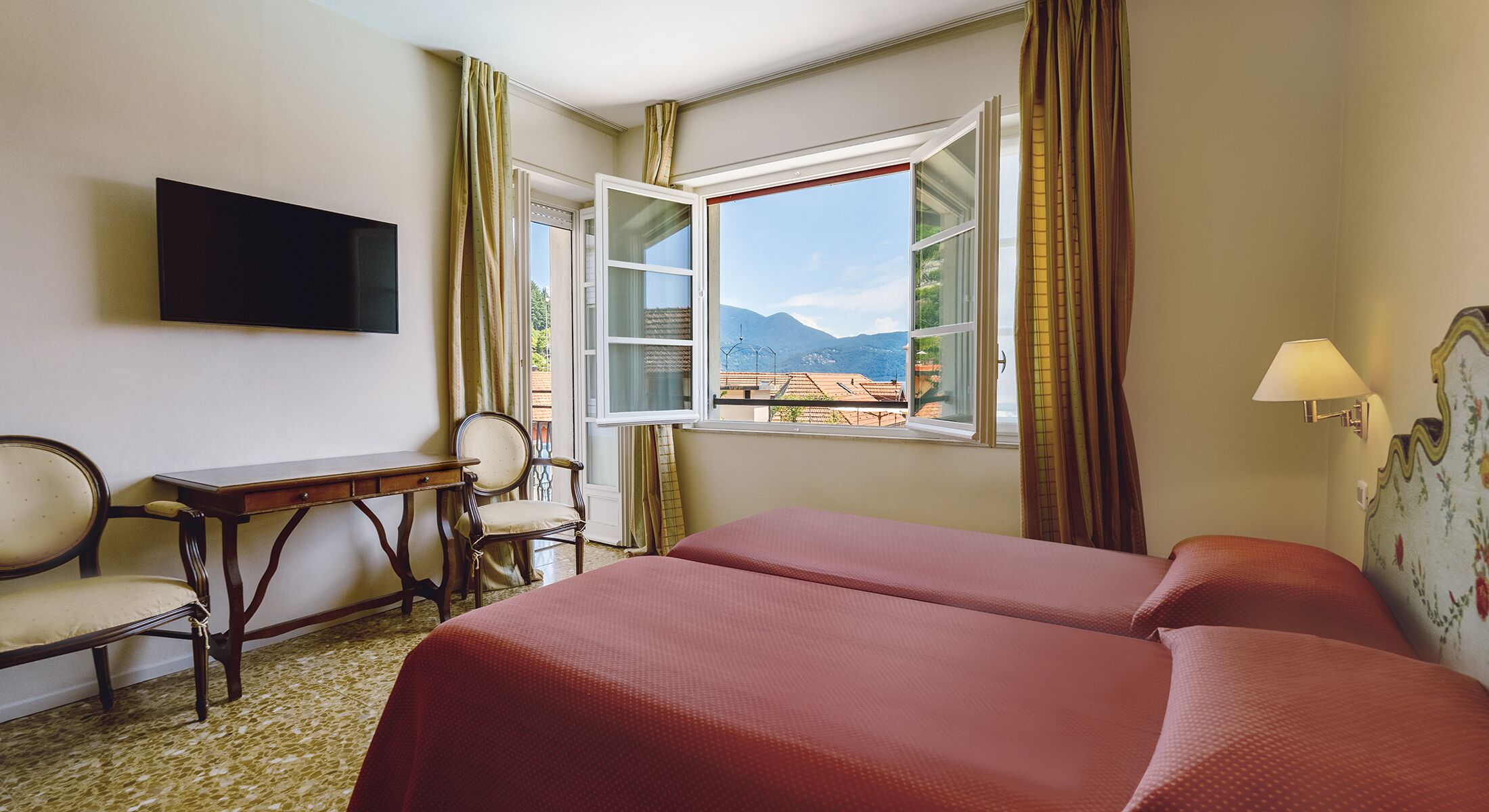Camera d'albergo con due letti, una scrivania, una sedia, una televisione e una finestra vista montagne.