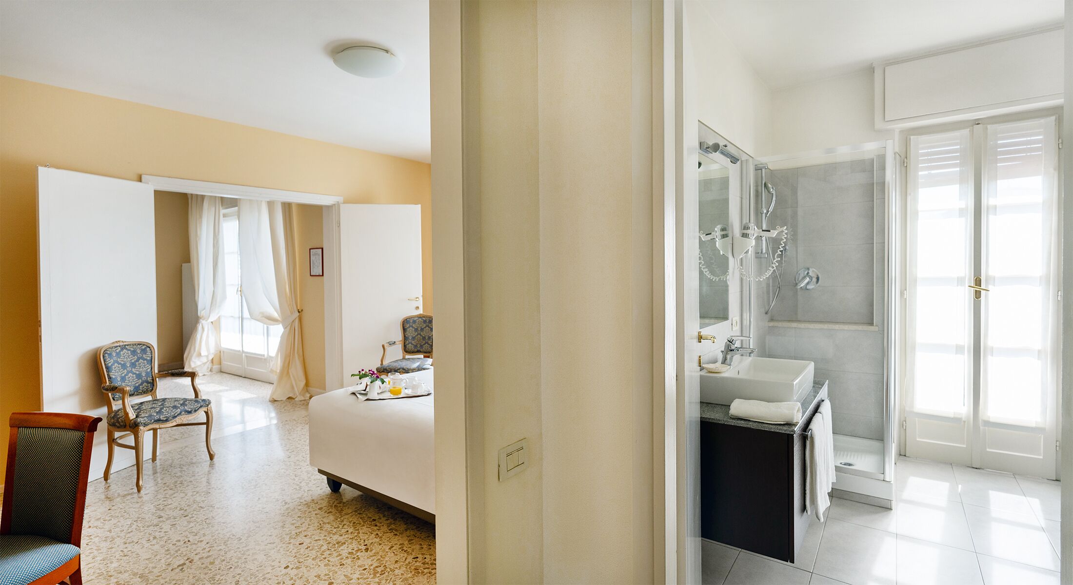 Una camera d'albergo con un letto, una sedia, un tavolino con un vassoio per la colazione e un bagno con lavandino e doccia.