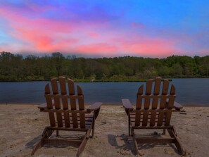 Two wooden Adirondack chairs facing a lake with a forested shoreline.