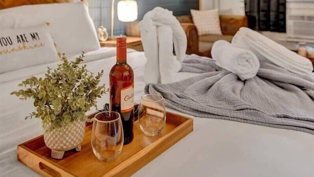 Et hotelværelse med en seng, et natbord med en flaske og glas, et bakke med en plante og glas, og udsigt til det fri.