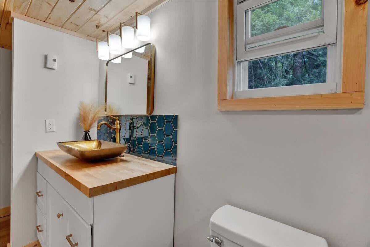 Une salle de bain avec un meuble-lavabo en bois, un pare-chocs en carreaux hexagonaux, un grand miroir et une fenêtre donnant sur les arbres.