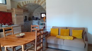A cozy living room with a wooden dining table, a sofa with yellow cushions, and a kitchen area with a stone archway. - Gîte Dans Maison en Pierre, Avec Piscine, à la Campagne, Proche Villefranche (Savignac)