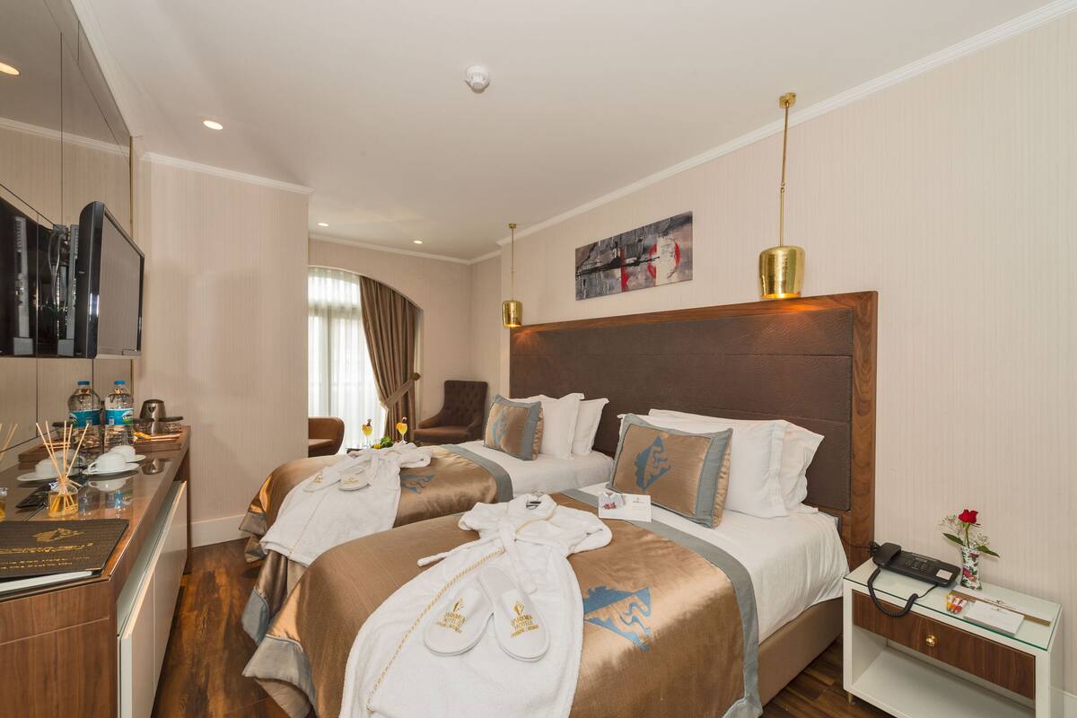 A hotel room with two beds, a desk with a flat-screen TV, a minibar, and a bathroom visible through a glass door.